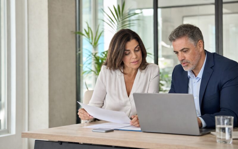 Attraktive realistische Berufstätige besprechen Altersteilzeit und Arbeitszeitplanung an einem Laptop in einem hellen Büro